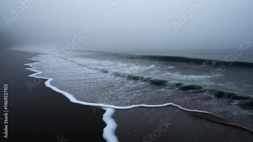 White waves gently lapping on a dark beach on a foggy day. The scene is moody and atmospheric, with a muted color palette of grays and dark browns. The focus is on the texture of the waves and the