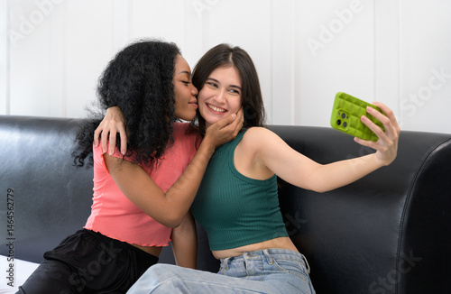 Two young women taking selfie tegether on a couch at home. African female in pink top kissing on cheek of Caucasian woman in green top. Love and friendship concept