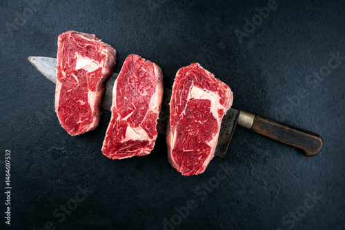 Dry aged raw beef entrecote cuts with fat marbling offered as top view on a black design board on a large kitchen knife