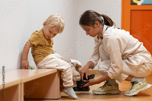 A mother assists her young child with putting on shoes as the child sits on a bench in a kindergarten or daycare hallway. Focus on daily routines, parental care, and getting ready.