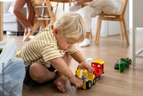 Fotografi A young blonde child sits barefoot on a wooden floor, deeply engrossed in playing with colorful toy trucks