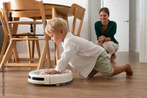 A young blonde child curiously interacts with a robotic vacuum cleaner on the wooden floor at home. The mother observes with a smile in the background, a modern family moment.