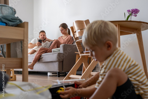 A young child sits on the floor engrossed in playing with toys, while in the background, parents relax together on the sofa, occupied with devices. Scene of modern family life and independent play.