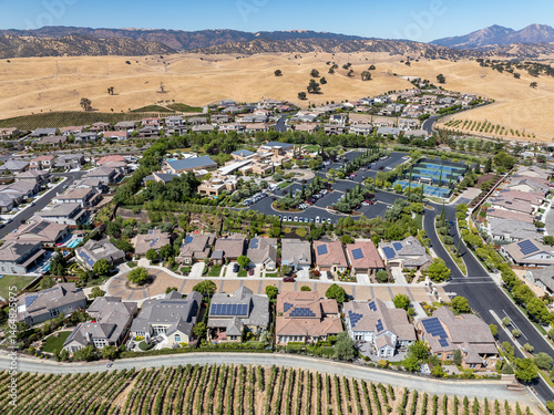 Aerial view of a suburban neighborhood with solar-paneled homes, swimming pools, and scenic rolling hills under clear blue skies in Antioch, California. Captured during summer.