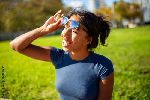 portrait of a young woman smiling and adjusting her sunglasses while sitting in a park