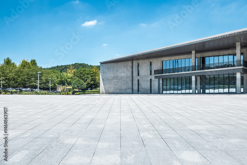 Empty stone square floor in front of a modern building exterior with green trees under a blue sky.