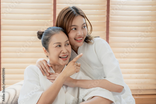 Teenage girl hugs senior mother joyfully while pointing forward with bright expressions and joyful connection at home. beauty routine concept, fresh radiant youthful look, feminine vibe photo