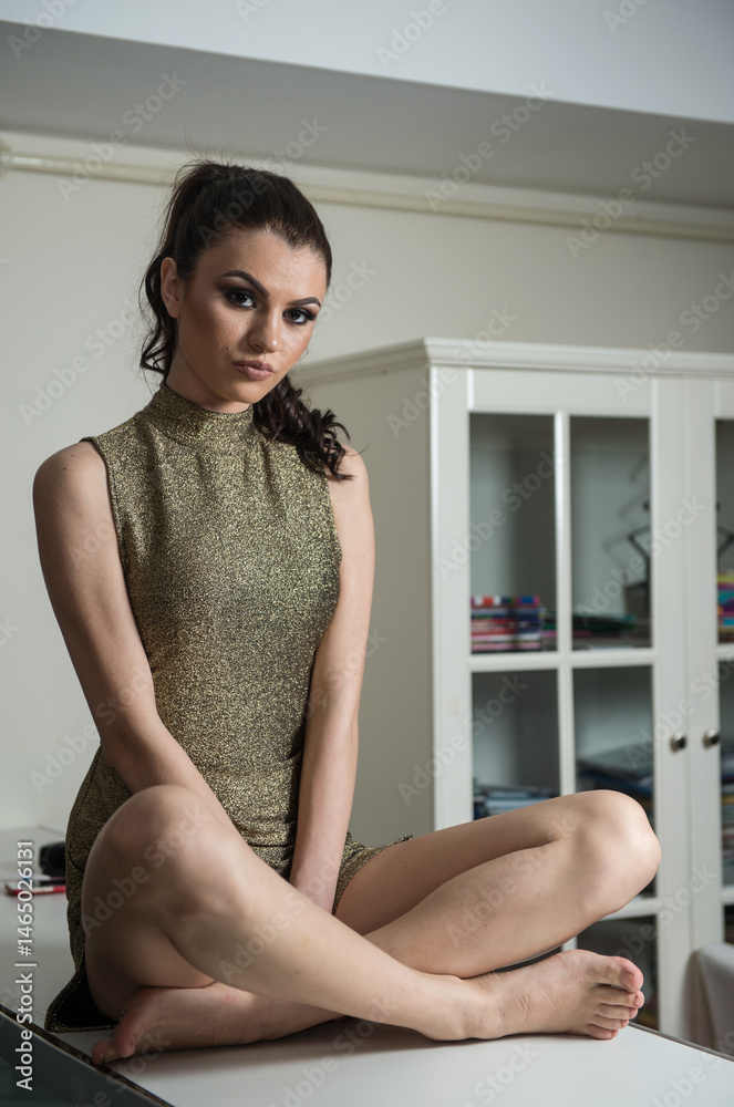 young dressed fashionable woman is sitting at a desk, Beautiful young woman in a gold short dress and high heels sitting on the table and posing