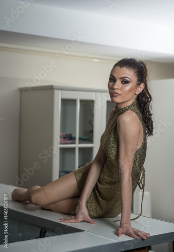 Obraz na płótnie young dressed fashionable woman is sitting at a desk, Beautiful young woman in a gold short dress and high heels sitting on the table and posing w salonie