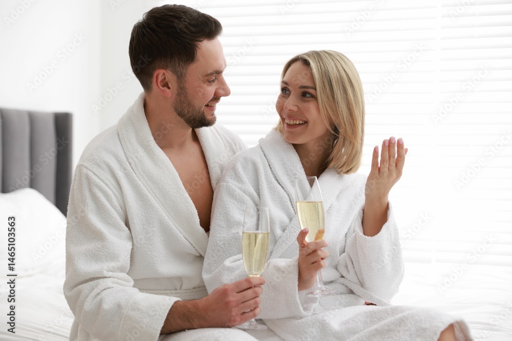Spa. Happy couple in bathrobes with glasses of champagne sitting on bed indoors Spa. Happy couple in bathrobes with glasses of champagne sitting on bed indoors
