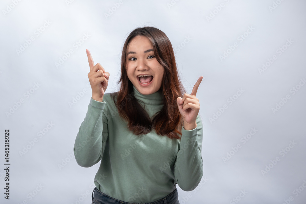 Asian woman smiling joyfully with mouth open, pointing both index fingers upward in a cheerful gesture, wearing green long-sleeve top against plain light background Asian woman smiling joyfully with mouth open, pointing both index fingers upward in a cheerful gesture, wearing green long-sleeve top against plain light background
