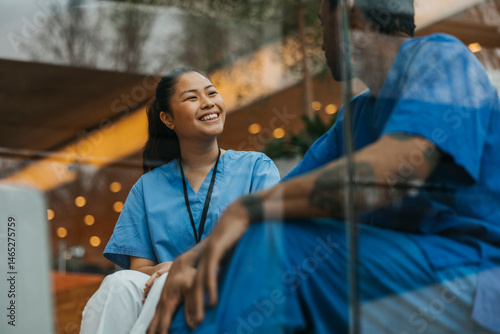Happy female healthcare professional talking with male coworker while sitting in hospital building seen through glass photo