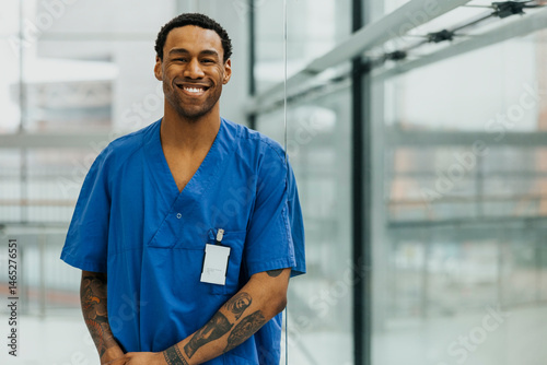 Portrait of happy male healthcare expert wearing medical scrubs with tattooed arm near glass wall