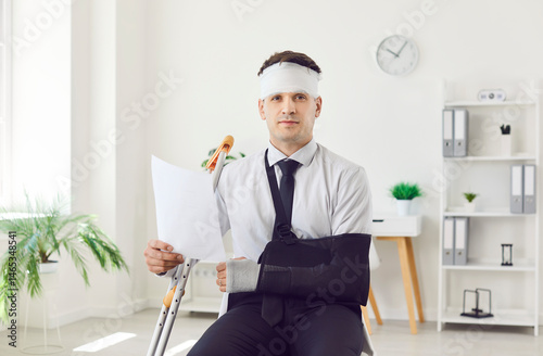 Portrait of a young handicapped injured man with broken arm and bandaged head after accident working in office holding document in hands and looking at camera. Insurance and compensation concept.