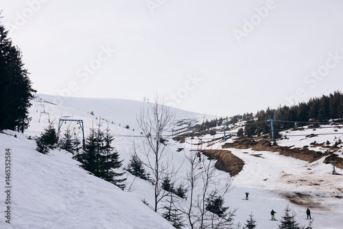 Partially snow-covered ski slope with ski lift under a cloudy sky.