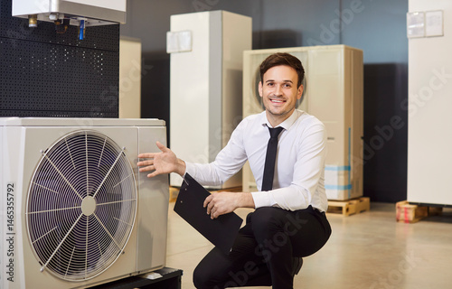 Happy, smiling man shop assistant with clipboard offers modern efficient sustainable ecologically friendly heat pump and HVAC split system for air conditioning available at their home appliances store