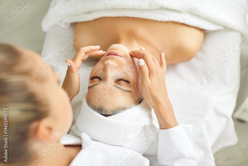Cosmetologist applies fabric nourishing mask to woman's face for rejuvenation and elimination of defects. Female client lying on massage table in spa. Concept of cosmetic procedures for women.