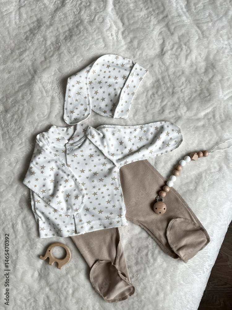 Scandinavian style in children's fashion is a set of neutral beige children's clothes for discharge. Baby's turtleneck, pants, cap, elephant teether, beads on a white plaid. Top view