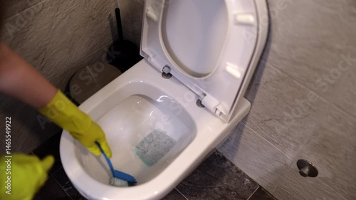 Woman wearing yellow gloves scrubs the inside of a toilet bowl with a cleaning brush. The bathroom features modern tiles and accessories. This routine promotes cleanliness and hygiene.