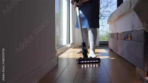 A woman is vacuuming a bright bedroom filled with natural light. Sunlight streams through the windows, illuminating the clean hardwood floors and creating a welcoming atmosphere.