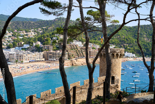 Scenic view of Tossa de Mar, with the Mediterranean Sea, a stone tower, trees, and a picturesque beach with boats.