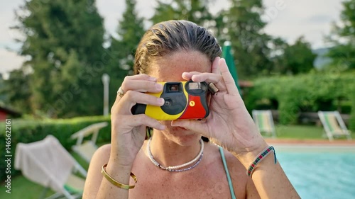 Young woman taking pictures with disposable stills camera in pool party outdoors. Portrait of happy cheerful female in fun party smiling takes film photo towards camera in garden with sun umbrellas