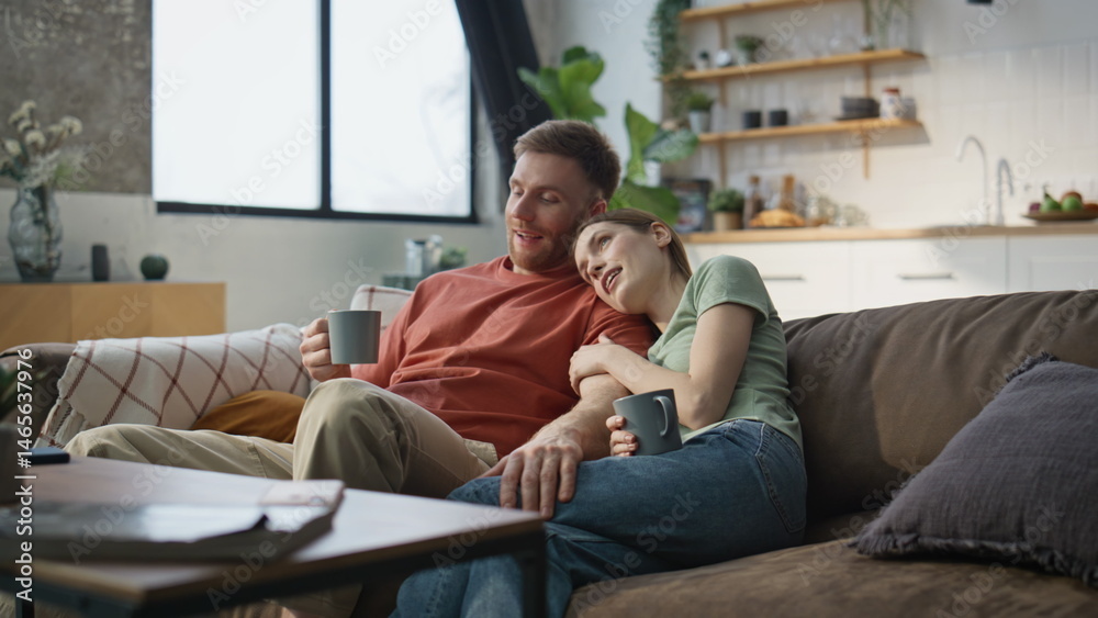 Happy lovers embracing sofa drinking morning coffee together. Woman cuddling man