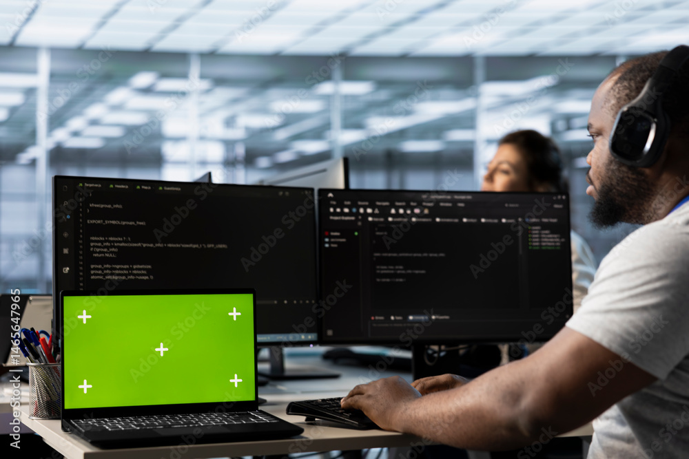 Isolated screen laptop next to man coding in server farm while listening music in headphones. Worker safeguarding hardware against unauthorized access with chroma key notebook, hearing songs