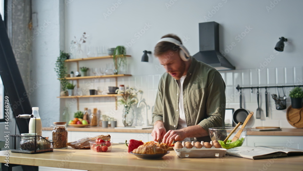 Singing man chopping pepper in kitchen apartment wearing earphones. Cheerful guy
