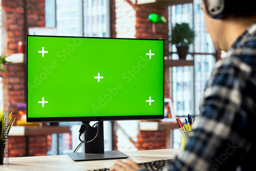 Worker listening music, analyzing data on green screen PC at desk, compiling analytical reports. Close up of employee wearing headphones using mockup computer, finishing project
