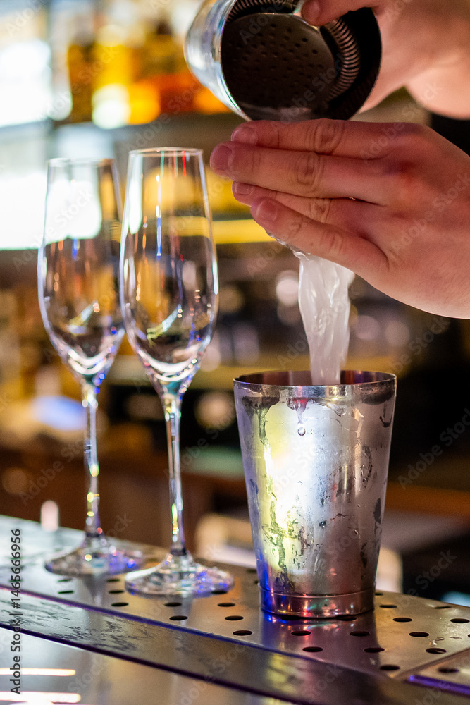 A bartender skillfully pours a cocktail from a shaker into a metal cup, set against a vibrant bar backdrop with champagne glasses and colorful lights.