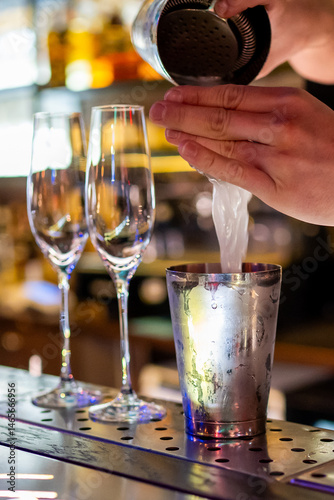 Obraz na płótnie A bartender skillfully pours a cocktail from a shaker into a metal cup, set against a vibrant bar backdrop with champagne glasses and colorful lights. w salonie
