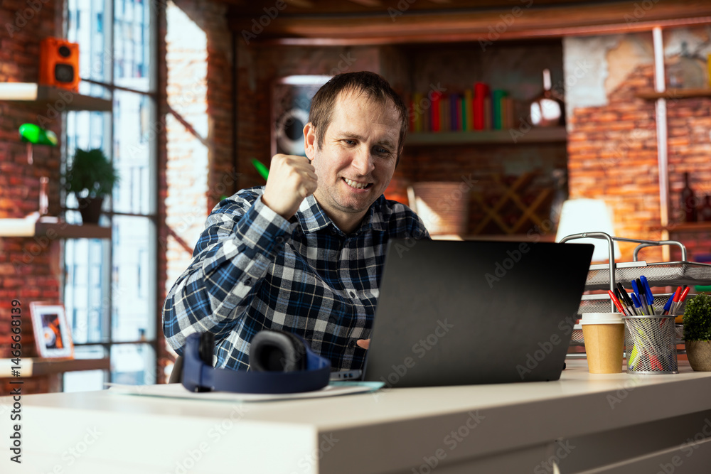 Happy worker at desk facing laptop screen, shaking fist to celebrate completion of task. Delighted teleworker in home office doing positive hand gesturing, looking at notebook screen