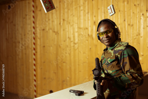 Portrait of smiling soldier in uniform doing training in shooting range, learning to use rifle. Happy military unit in firing range practicing with combat weapons, wearing protective equipment