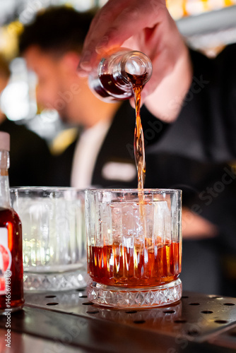 A bartender skillfully pours dark liquor into a glass with ice cubes, capturing the essence of mixology and craftsmanship in a vibrant bar setting.