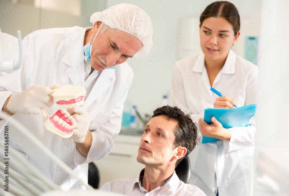 During appointment, mature man dentist and girl nurse communicates with male patient, shows model of jaws and explains mechanism of defect formation. During appointment, mature man dentist and girl nurse communicates with male patient, shows model of jaws and explains mechanism of defect formation.