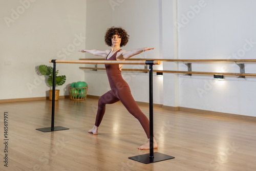 Confident woman in elegant activewear training arms and posture using a ballet barre in spacious studio with wood floor, soft light and indoor plant. Horizontal, copy-space