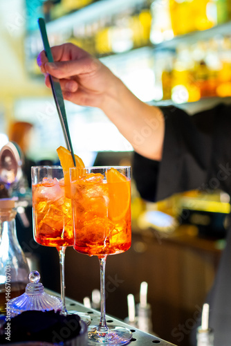 Obraz na płótnie A bartender prepares Aperol Spritz cocktails with orange slices, creating a vibrant and refreshing scene at a bar. The bright colors contrast with ice cubes, enhancing the visual appeal. w salonie