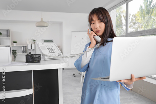Upset female Asian medical intern talking by telephone near reception in clinic