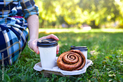 picnic food and drinks on the grass in a sunny park.two coffee cups and a large pastry, with a hand reaching for a cup