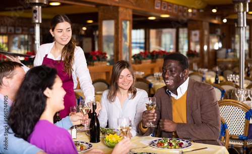 Young waitress serving bottle of wine to cheerful multiracial group of adults having friendly get-together in cozy restaurant..