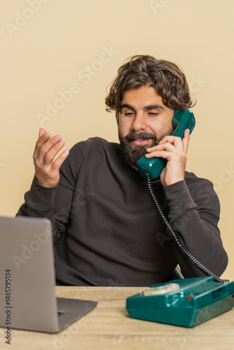 Happy bearded Hindu businessman sitting at table talking on wired telephone call conversation communication with client. Young freelancer Indian guy using laptop computer on beige studio background.