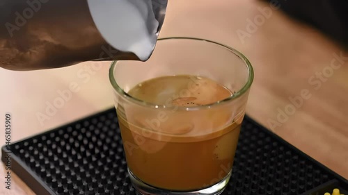 Pouring White Milk into Espresso Coffee in Clear Glass on Black Mat Placed on a Wooden Table Creating Latte Art in Bright Indoor Light