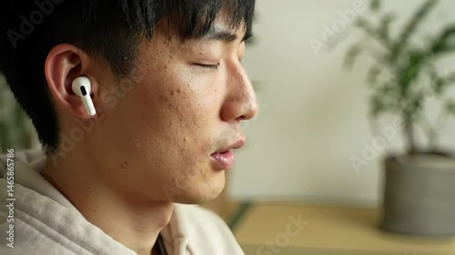 Sweaty Man with Eyes Closed Meditating Indoors Wearing Wireless Earphones in Natural Light with a Potted Plant on a Tatami Mat