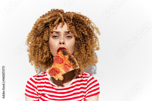 Obraz na płótnie Young woman with curly hair enjoying a slice of pizza, expressing joy and indulgence, wearing a striped shirt, minimalist white background enhancing the vibrant colors w salonie