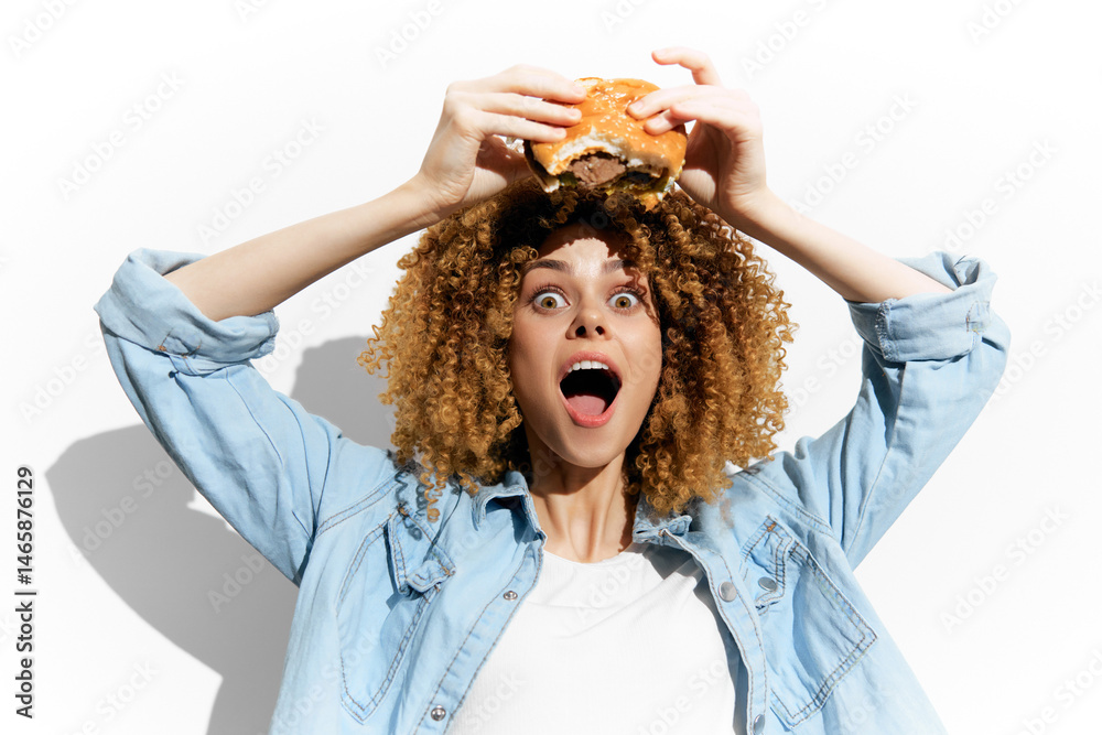 Enthusiastic young woman with curly hair joyfully holding a burger, showcasing excitement for fast food against a white background, perfect for food related content