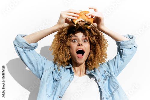 Obraz na płótnie Enthusiastic young woman with curly hair joyfully holding a burger, showcasing excitement for fast food against a white background, perfect for food related content w salonie