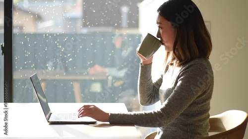Young Woman in Gray Shirt Works at Laptop with Coffee Cup at Bright Desk With Geometric Window Light and Another Person in Background