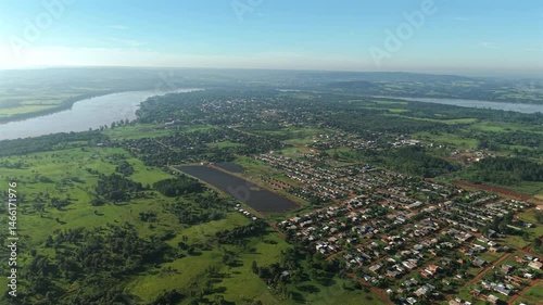 Wide aerial perspective shows the residential layout of San Javier, Misiones, Argentina, surrounded by green fields, a solar panel facility, Uruguay River curving along the border, drone shot