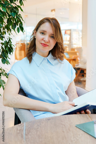 Portrait of a confident businesswoman sitting at a table in a modern office or coworking space. She is smiling warmly and looking at the camera while holding an open notebook. 